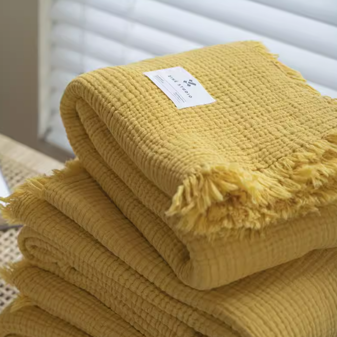 Yellow organic cotton waffle textured fringe throw blanket folded neatly showing soft gauze weave and decorative fringe edges.