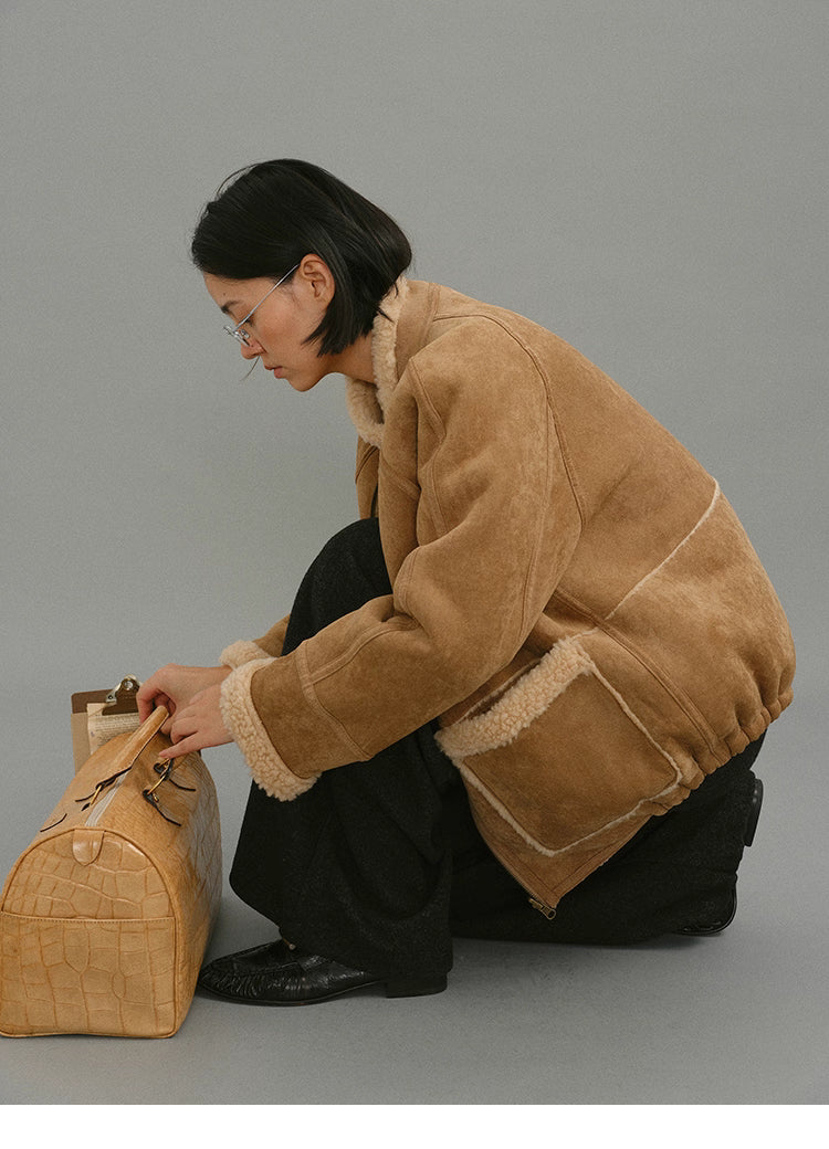 Model wearing light camel suede shearling jacket kneeling beside travel bag