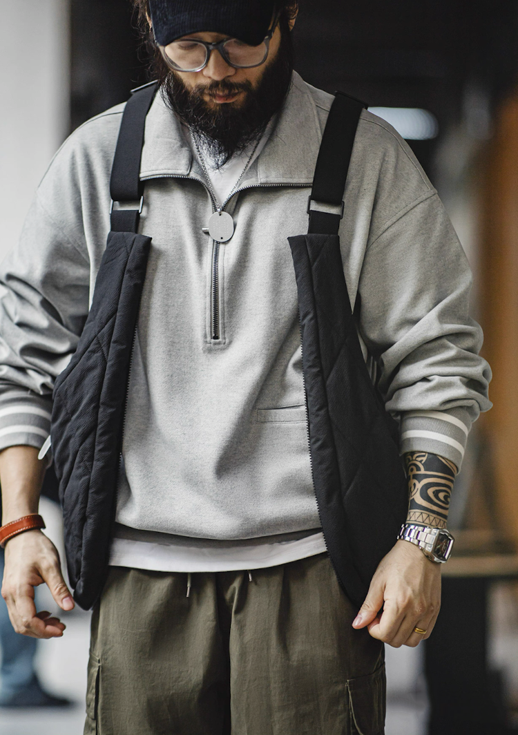 Model wearing black quilted convertible vest bag over gray zip-neck sweater urban style.