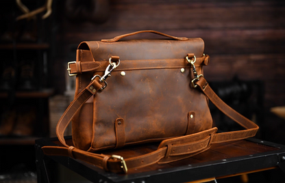 Rear view of leather satchel with shoulder strap and brass clips on wooden desk.
