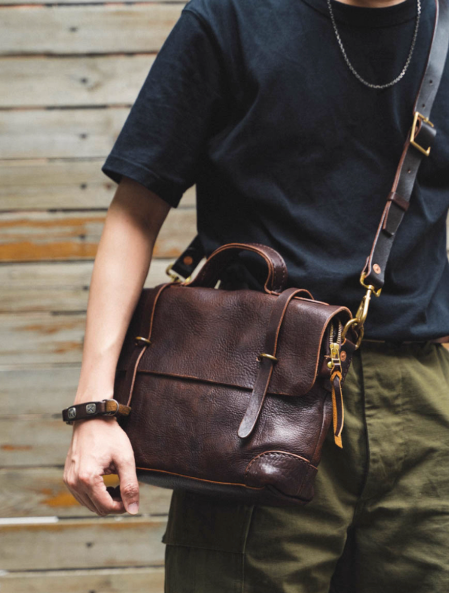 Person wearing dark t-shirt and khaki pants carrying dark brown full-grain leather messenger bag over shoulder outdoors