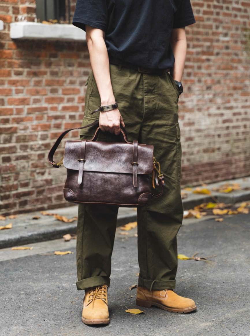 Model holding vintage brown leather messenger bag by handle while standing on city street in olive pants