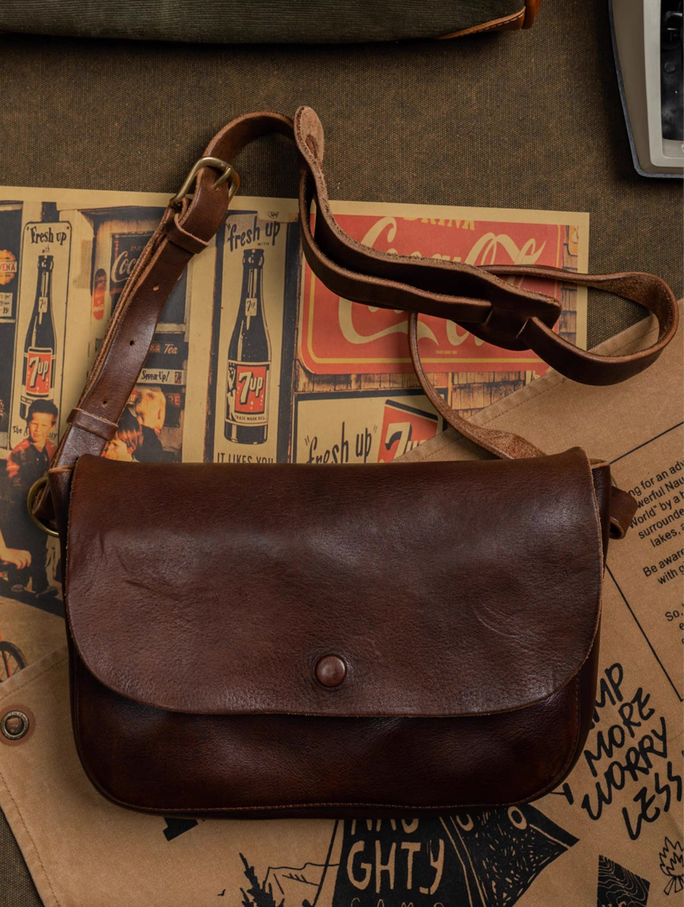 Dark brown full-grain leather crossbody bag laid flat on vintage paper backdrop with 7up posters