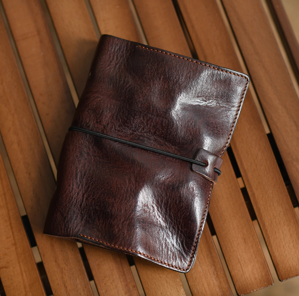 Dark brown full-grain leather journal with black elastic closure placed flat on wooden slatted surface.