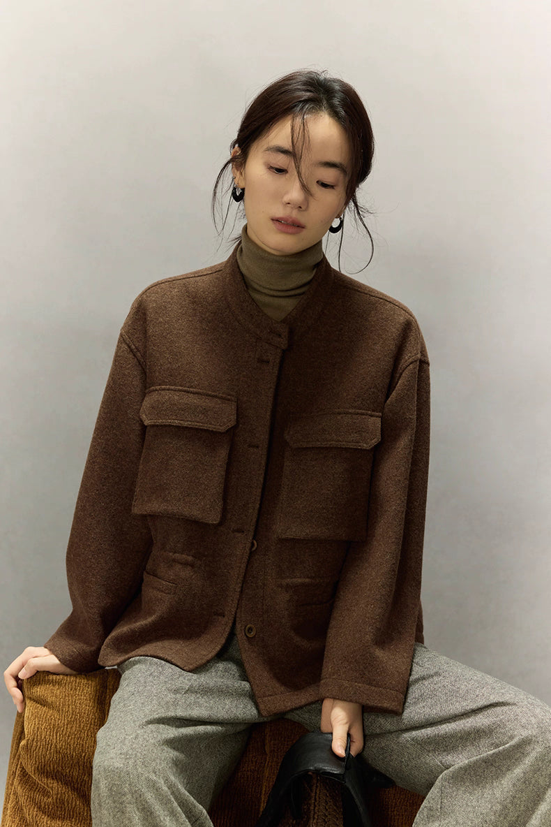 Woman seated wearing dark coffee alpaca wool field jacket and grey trousers, front view in soft studio light