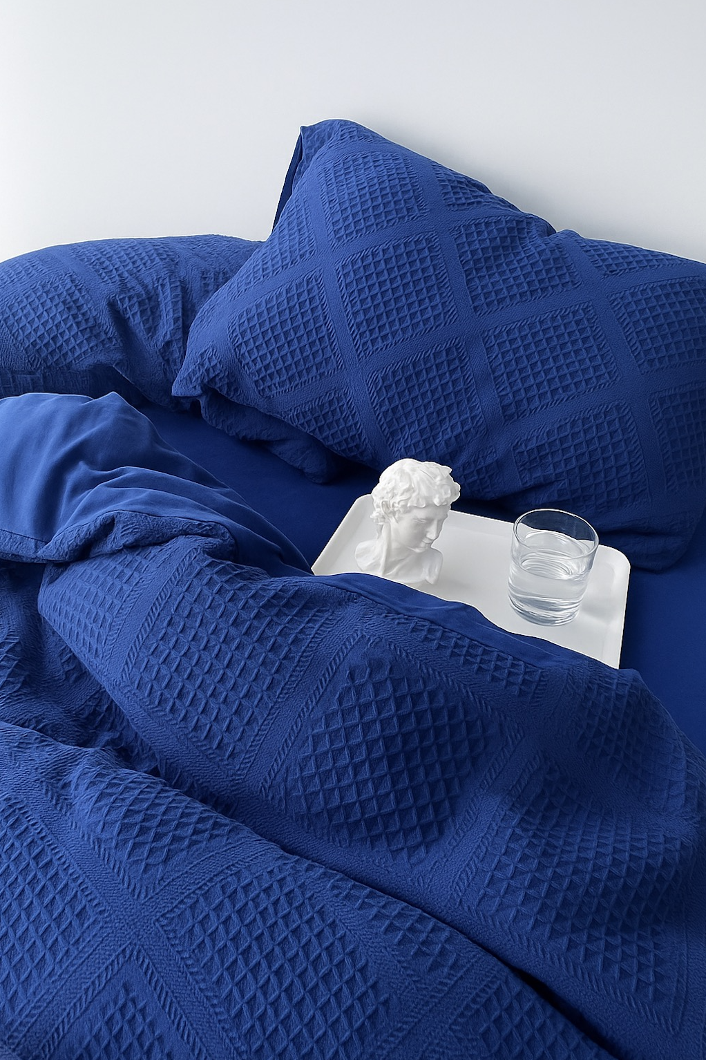 Extreme close-up of royal blue textured jacquard bedding with pillow; white bust and glass on bedside tray.