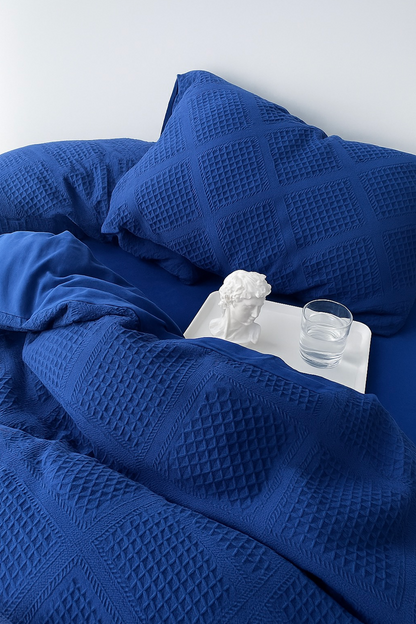 Extreme close-up of royal blue textured jacquard bedding with pillow; white bust and glass on bedside tray.