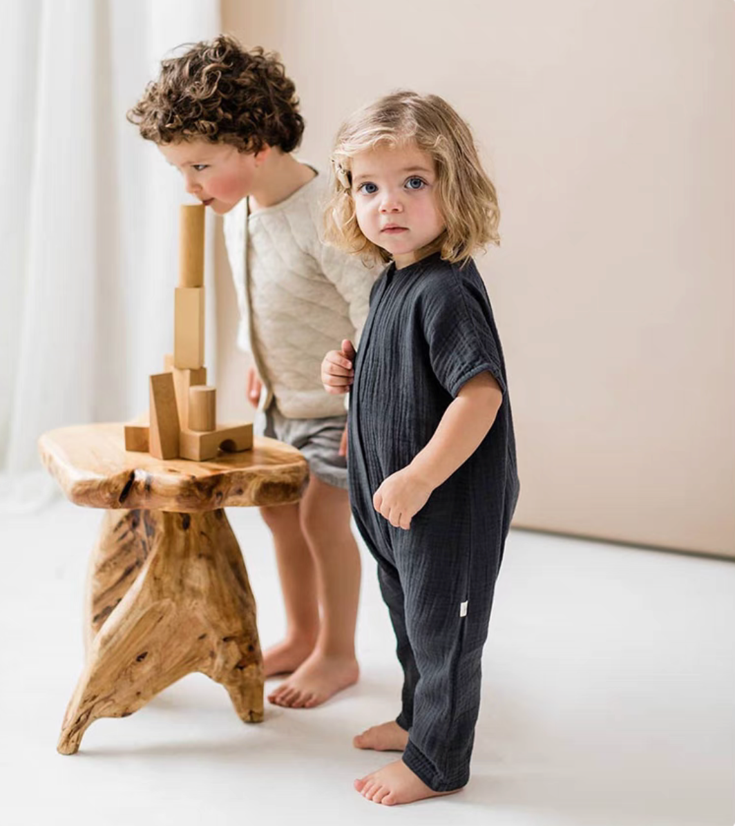 Toddler wearing charcoal gray cotton romper standing beside wooden table indoors
