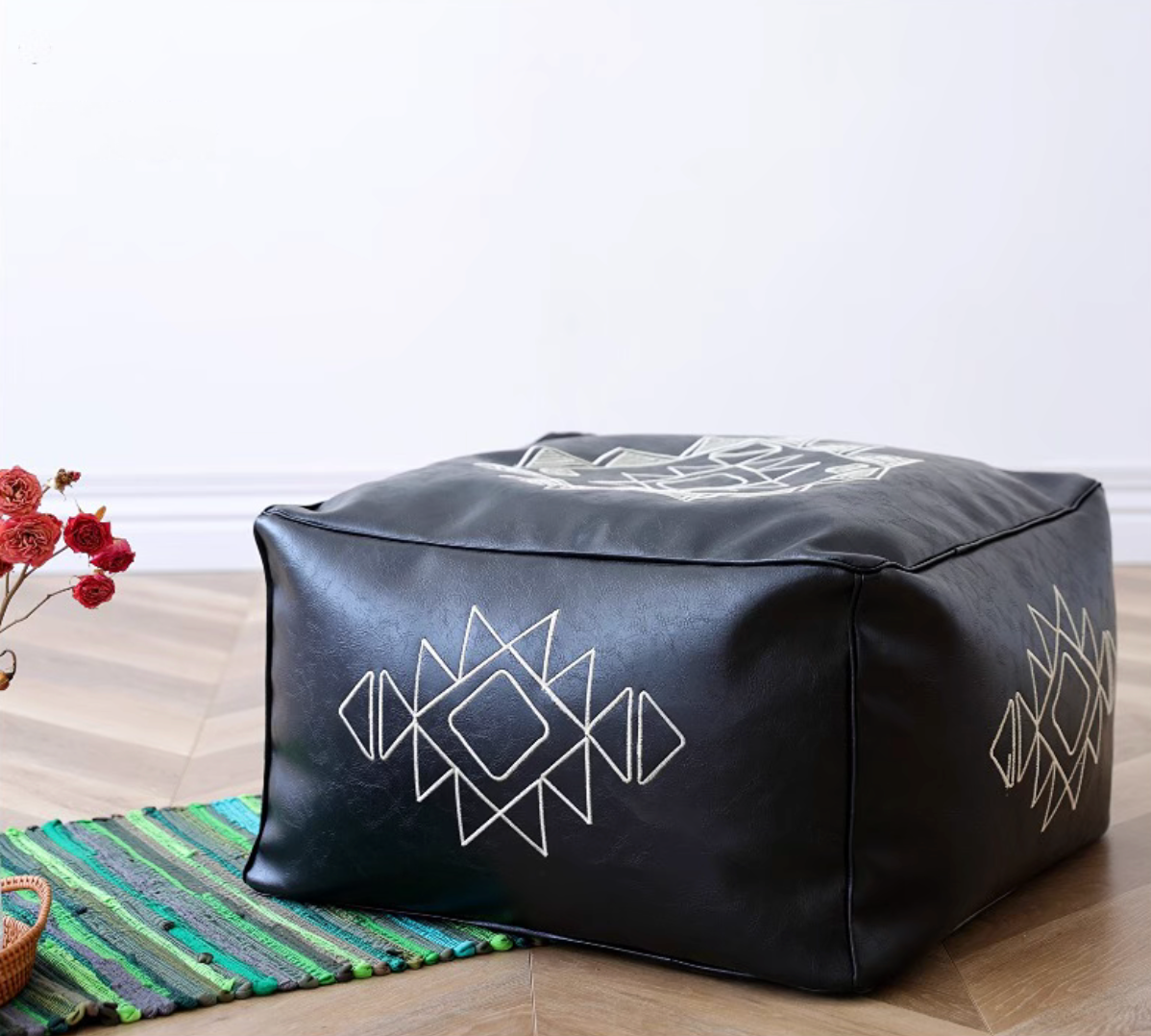 Black embroidered square bean bag sofa with white geometric pattern placed on a wooden floor with decorative rug and flowers