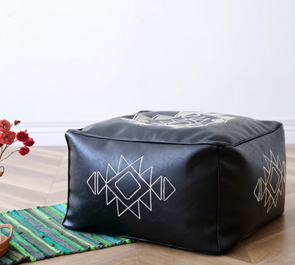 Black embroidered square bean bag sofa with white geometric pattern placed on a wooden floor with decorative rug and flowers