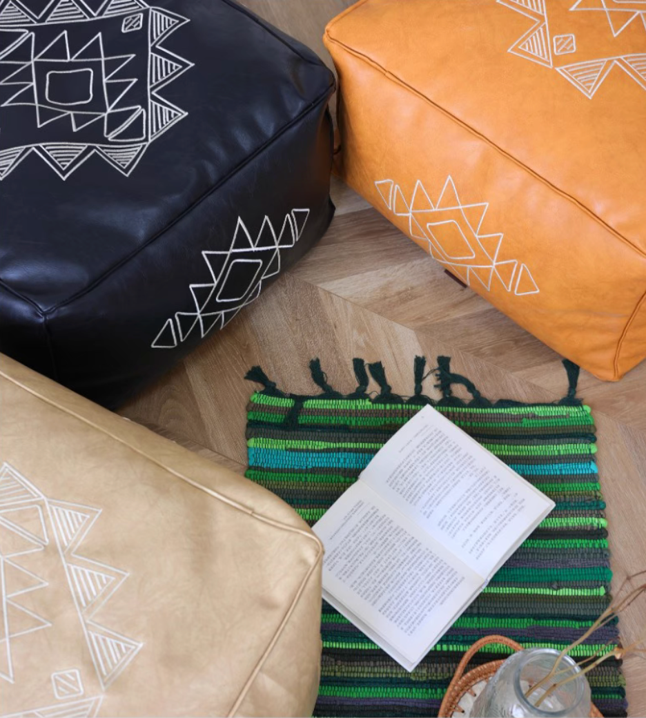 Top view of black, orange yellow, and khaki embroidered square bean bag sofas arranged on a floor with a green woven rug and open book