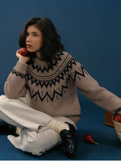 Woman sitting on the floor wearing a taupe and black Fair Isle merino wool sweater, holding a pear by her face against a blue background.
