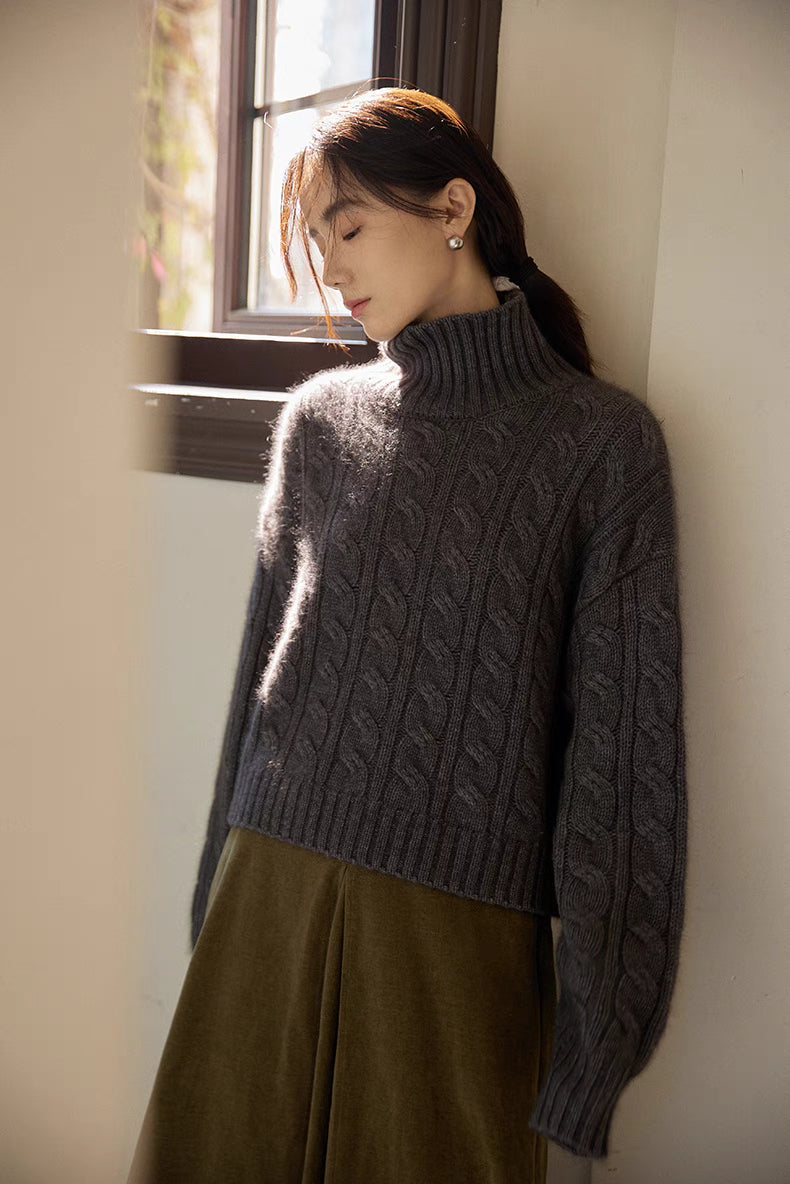 Dark gray cable-knit sweater worn while leaning by window highlighting soft drape and warm knit texture