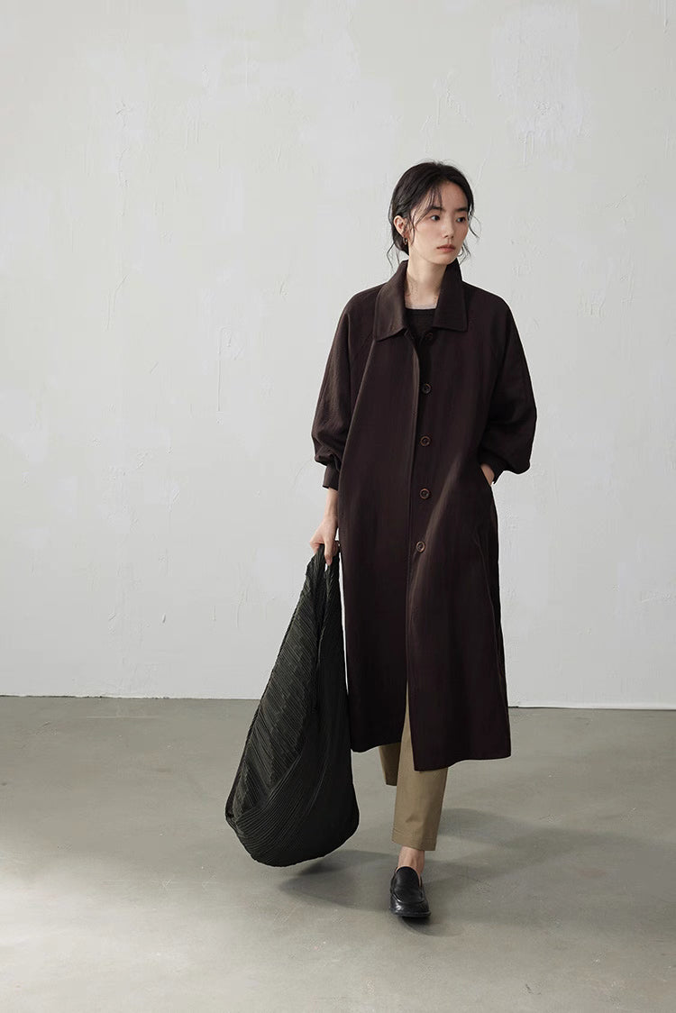 Model in a coffee brown fluid-drape Tencel trench coat, full front view, holding a large black tote bag.
