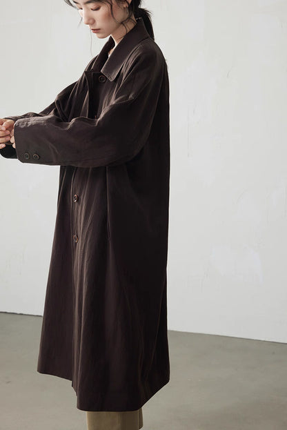 Side view of the coffee brown trench coat with model adjusting sleeve, showing relaxed raglan shoulder and cuff detail.