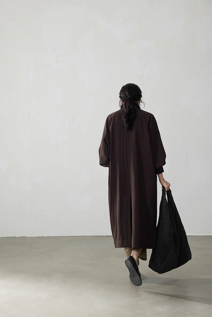 Full back view of the coffee brown fluid-drape Tencel trench coat as the model walks away holding a black bag.