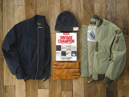 Two MA-1 jackets in navy and army green with vintage magazine, hat, and gloves flat lay.