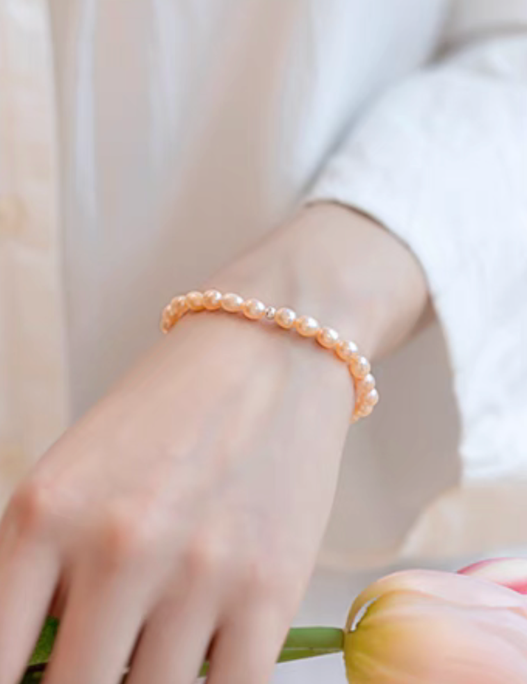 Lifestyle image of peachy pink freshwater pearl stretch bracelet worn on wrist with soft floral background.