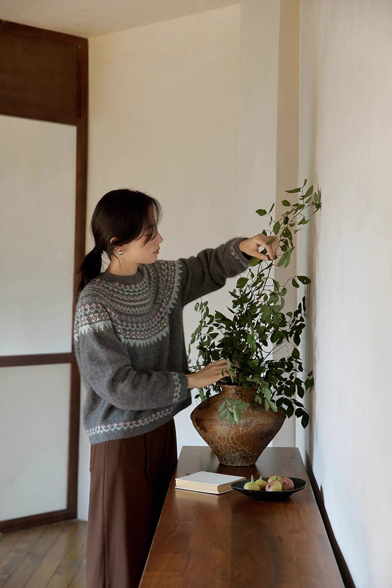 Model wearing pine gray Frosted Peaks Fair Isle Sweater arranging plant indoors highlighting cozy lifestyle and Nordic-inspired design