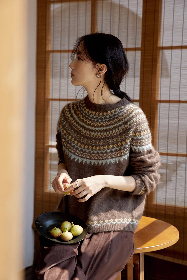 Model wearing Frosted Peaks Fair Isle Sweater in smoky brown sitting by wooden window in side profile showcasing Nordic pattern and soft knit texture