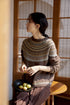 Model wearing Frosted Peaks Fair Isle Sweater in smoky brown sitting by wooden window in side profile showcasing Nordic pattern and soft knit texture