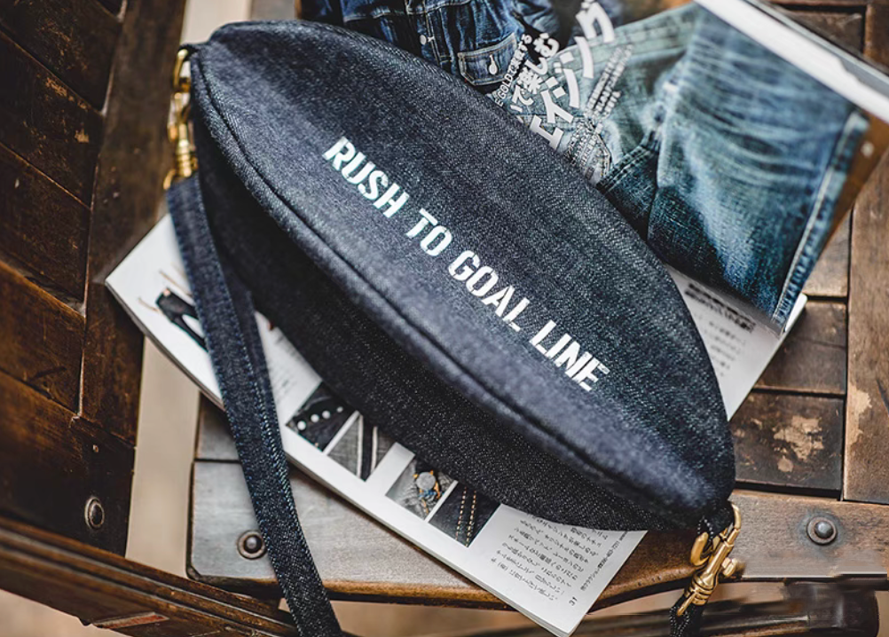 Goal-Line Denim Sling Crossbody Bag in denim blue flat lay on magazine showing rugby inspired oval shape premium denim texture bold printed text and brass hardware accents