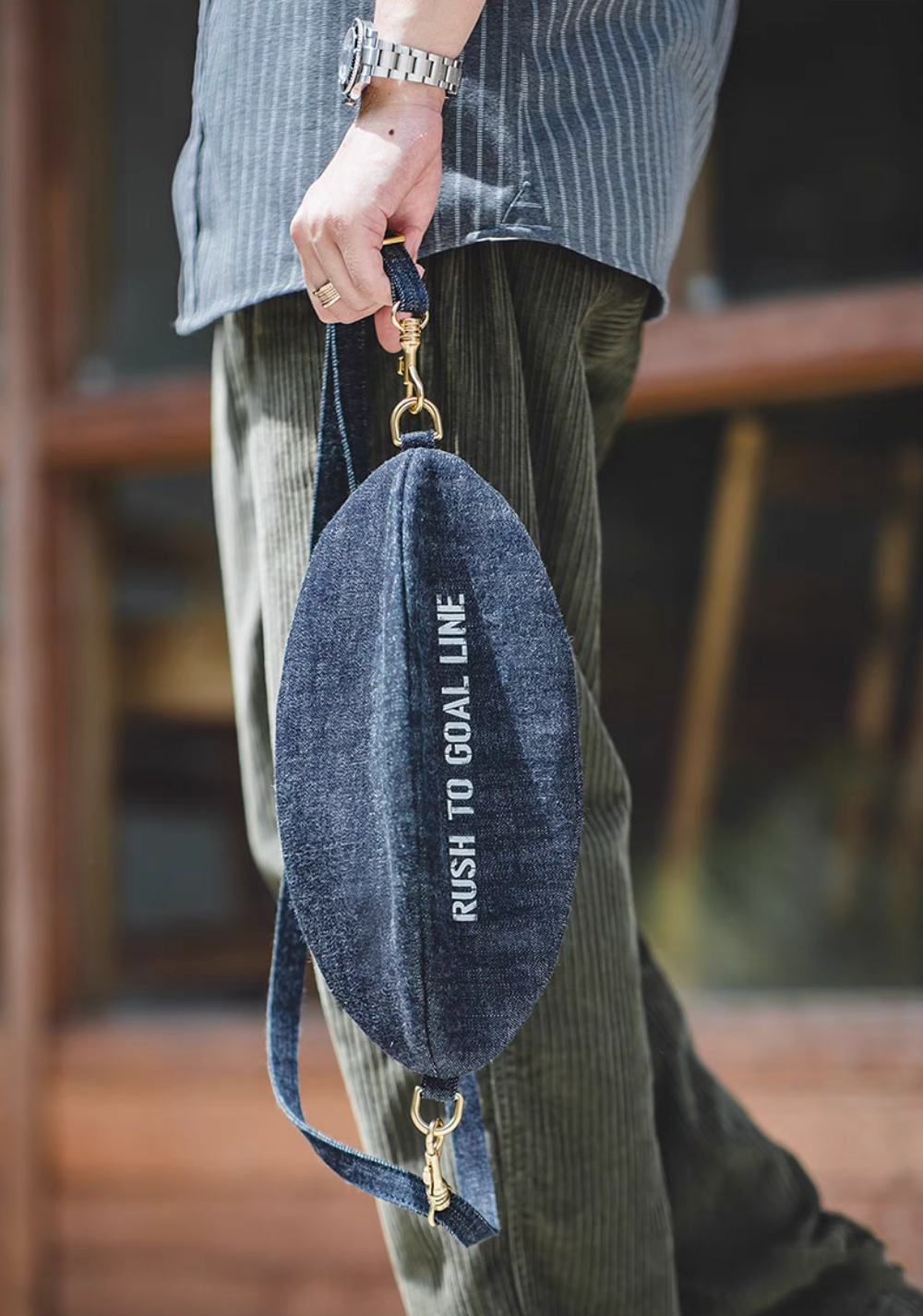 Goal-Line Denim Sling Crossbody Bag denim blue carried by hand showcasing durable strap brass clasp hardware and compact oval silhouette