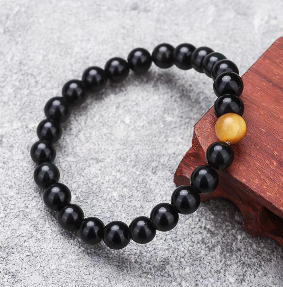Black tiger’s eye beaded bracelet featuring a single golden tiger’s eye accent bead shown on textured surface with wooden detail