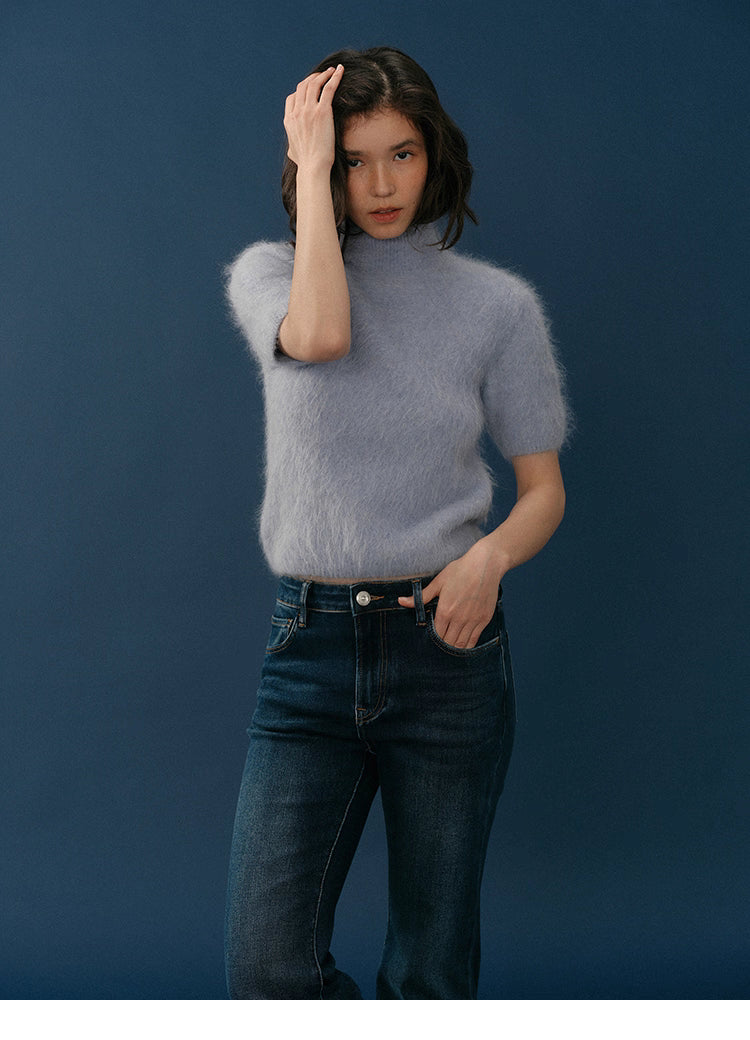Model wearing the light lavender HaloFuzz Alpaca Turtleneck Tee with dark denim, front view, one hand in her hair against a deep blue background.