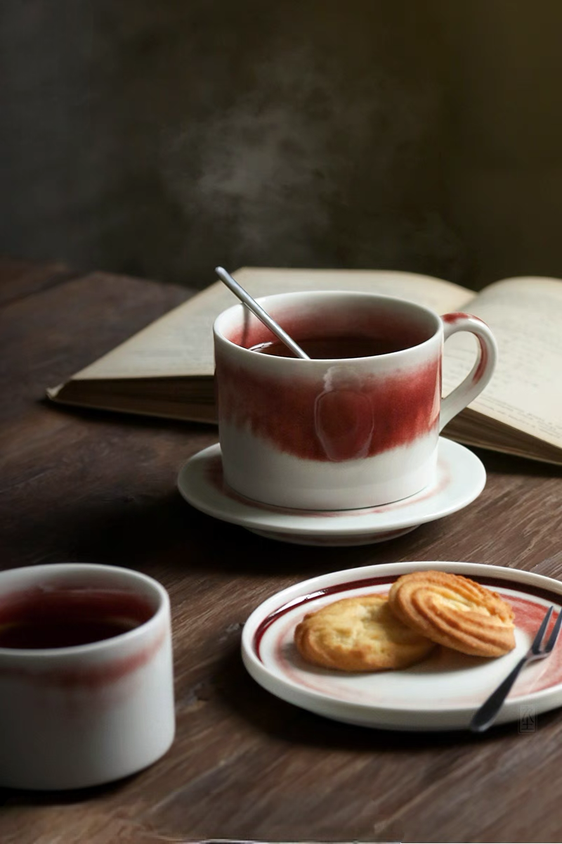 Beige and red handcrafted ceramic cup and saucer set used for tea with book and dessert creating a warm vintage coffee moment