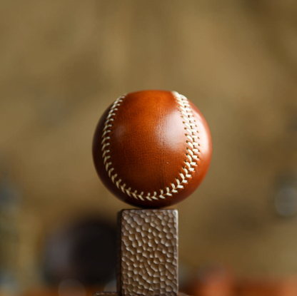 Hand-stitched caramel leather baseball balanced on textured stand with warm studio lighting.
