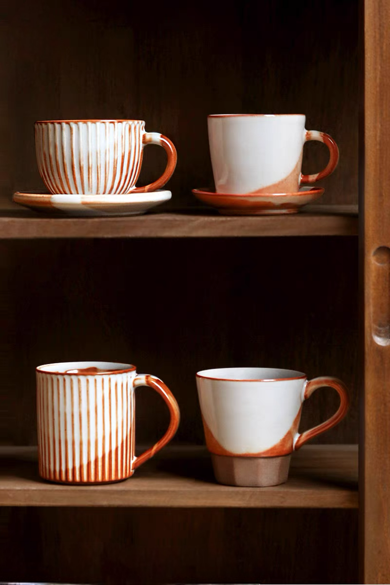 Off-white and orange handcrafted vintage ceramic mugs in multiple styles displayed on wooden shelf with varied shapes and textures