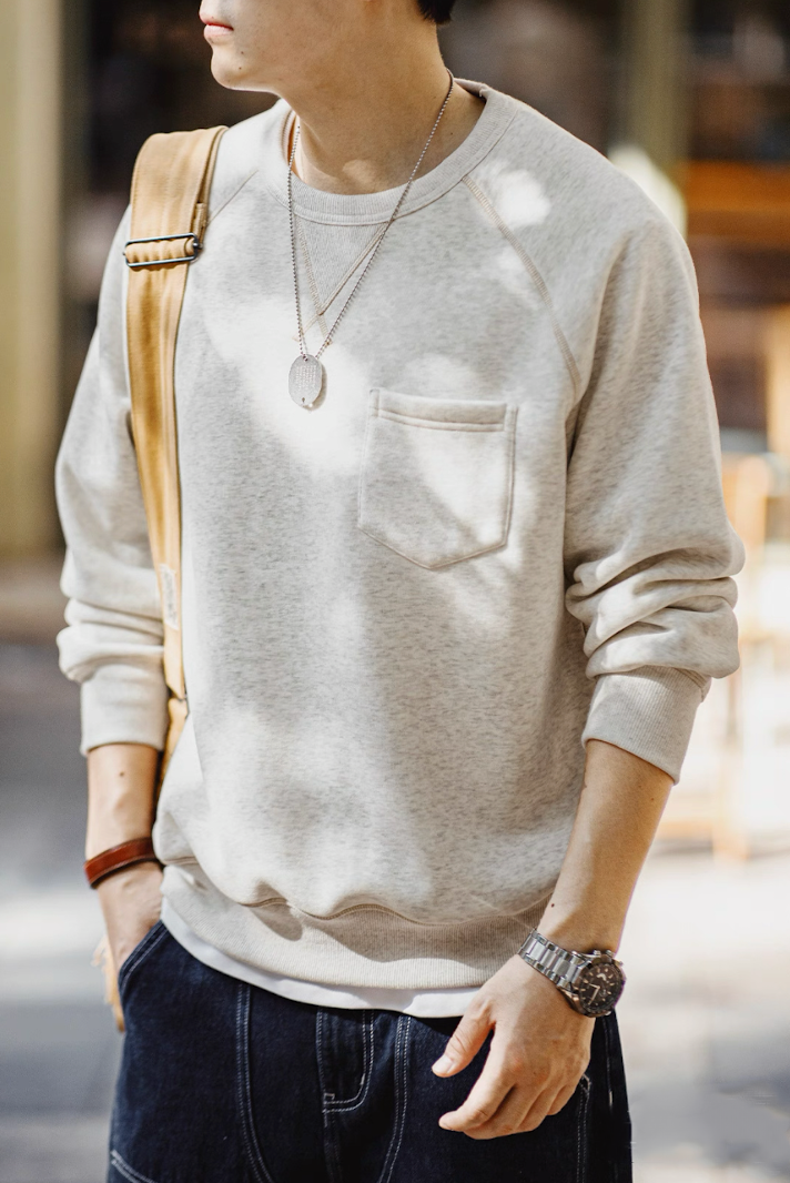 Model wearing light gray crewneck outdoors with denim and beige bag, showing chest pocket detail.