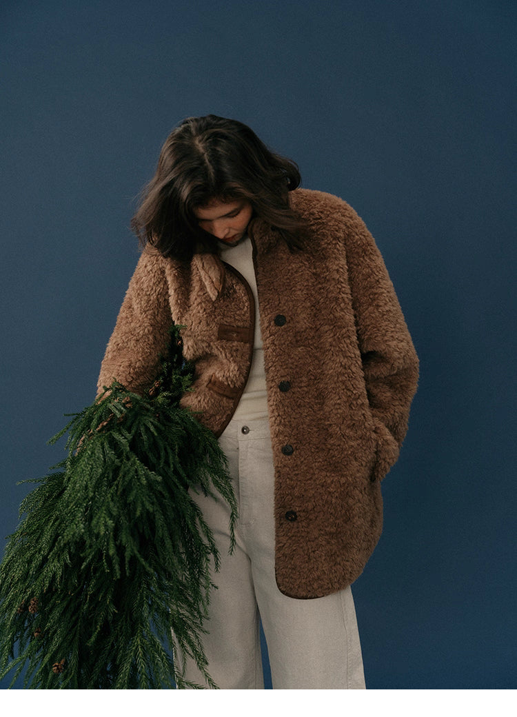Model looking down while wearing chocolate sherpa coat, holding winter greenery branches.