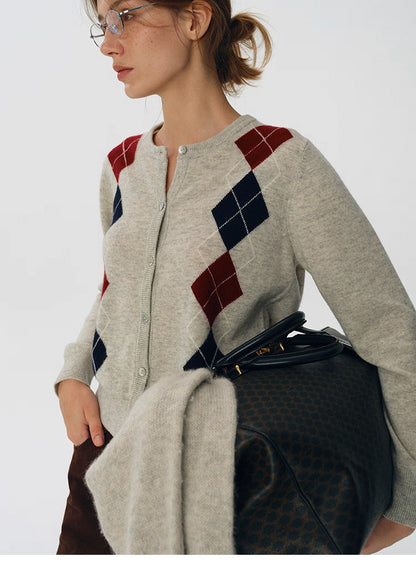 Side close-up of oat beige argyle wool cardigan, paired with a dark patterned travel bag and brown trousers.