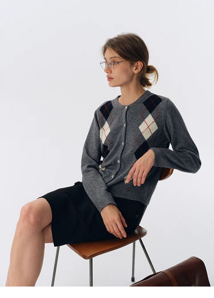Charcoal gray wool argyle cardigan worn while seated on a chair, paired with a black skirt and glasses.