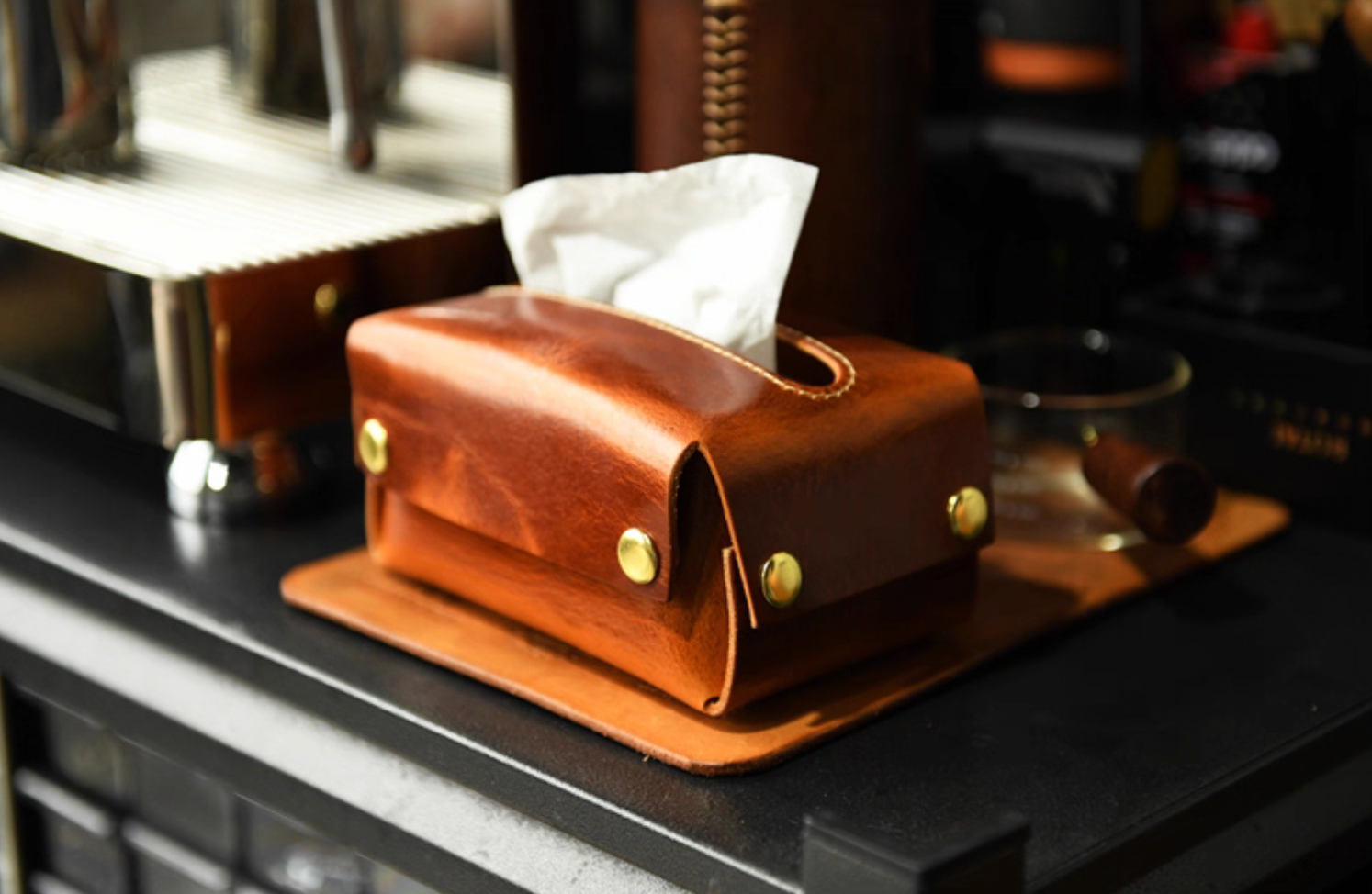 Handcrafted caramel leather tissue box on coffee counter beside glass cup and cigar.
