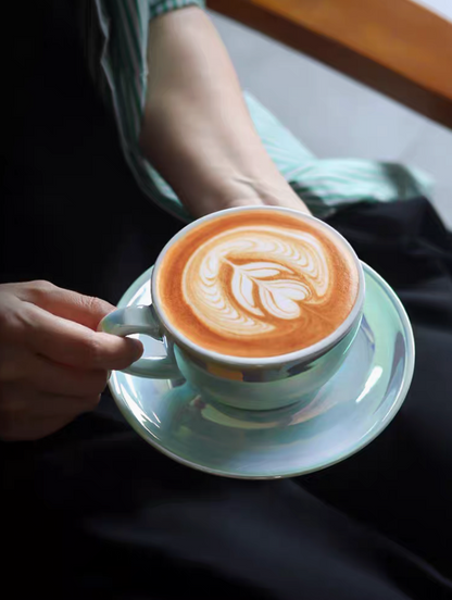 Luminous iridescent blue ceramic coffee cup and saucer with latte art held in hand creating a warm lifestyle coffee moment