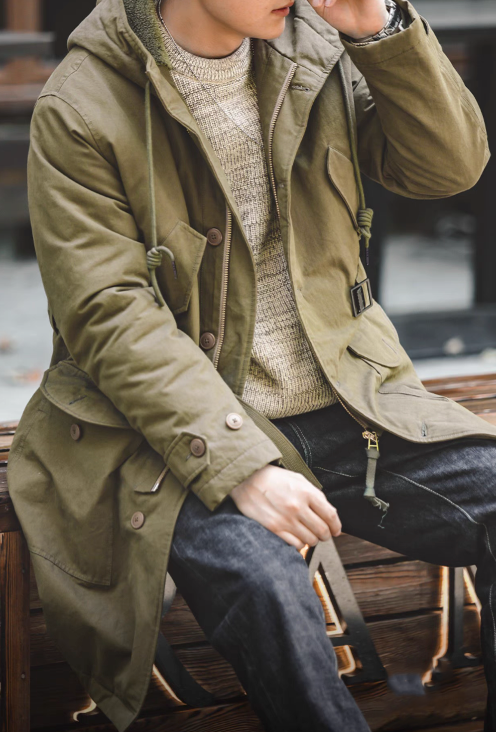 Model wearing M47 Heritage Explorer Parka Coat in military green seated view with open front layered over knitwear showing zipper and pocket details
