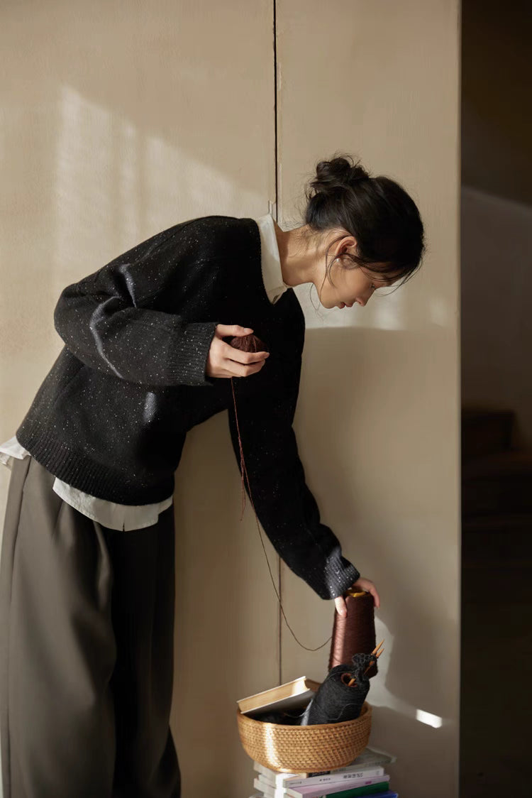 Black speckled merino wool sweater worn while reaching toward basket in warm light showing relaxed fit and textured knit