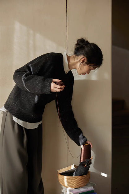 Black speckled merino wool sweater worn while reaching toward basket in warm light showing relaxed fit and textured knit