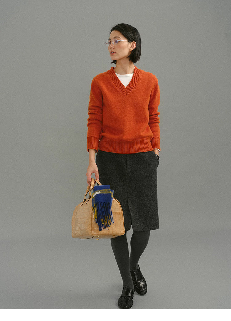 Model wearing a rust-orange Merino V-neck sweater styled with a charcoal knee-length skirt, grey tights, black loafers, and carrying a tan handbag with a blue plaid scarf.