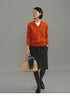 Model wearing a rust-orange Merino V-neck sweater styled with a charcoal knee-length skirt, grey tights, black loafers, and carrying a tan handbag with a blue plaid scarf.