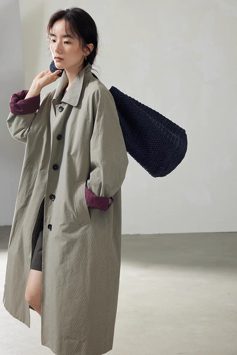 Light khaki gray cotton trench coat styled with navy textured shoulder bag, showing relaxed everyday look.