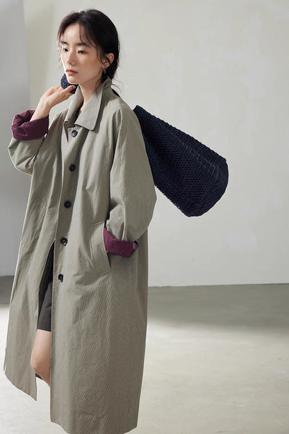 Light khaki gray cotton trench coat styled with navy textured shoulder bag, showing relaxed everyday look.
