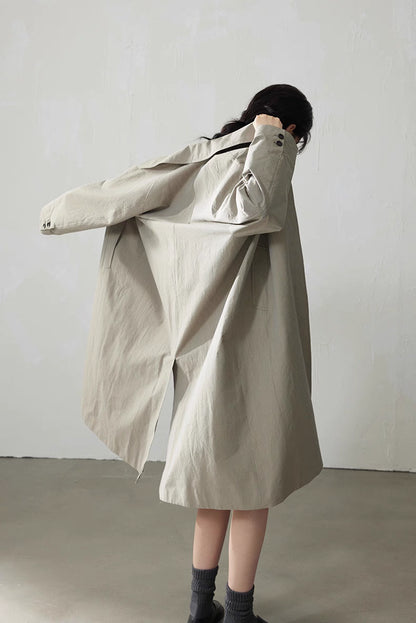Back view of light khaki gray trench coat swinging as the model puts it on, showing fluid straight silhouette.