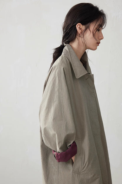 Side profile of model in light khaki gray trench coat with rolled burgundy cuff and relaxed sleeve shape.