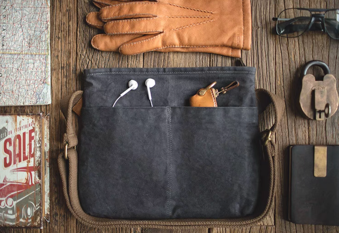 Dark grey waxed canvas crossbody bag with front pockets holding earphones and a leather key holder, styled with gloves, map, and everyday carry items on a wooden surface.