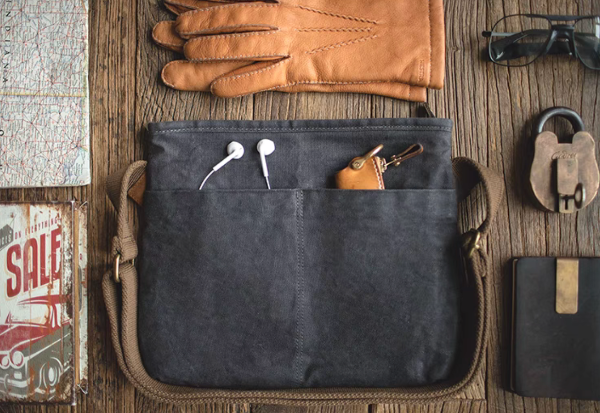 Dark grey waxed canvas crossbody bag with front pockets holding earphones and a leather key holder, styled with gloves, map, and everyday carry items on a wooden surface.