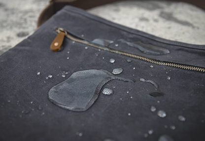 Dark grey waxed canvas crossbody bag surface with water droplets demonstrating water-resistant material and zipper detail.